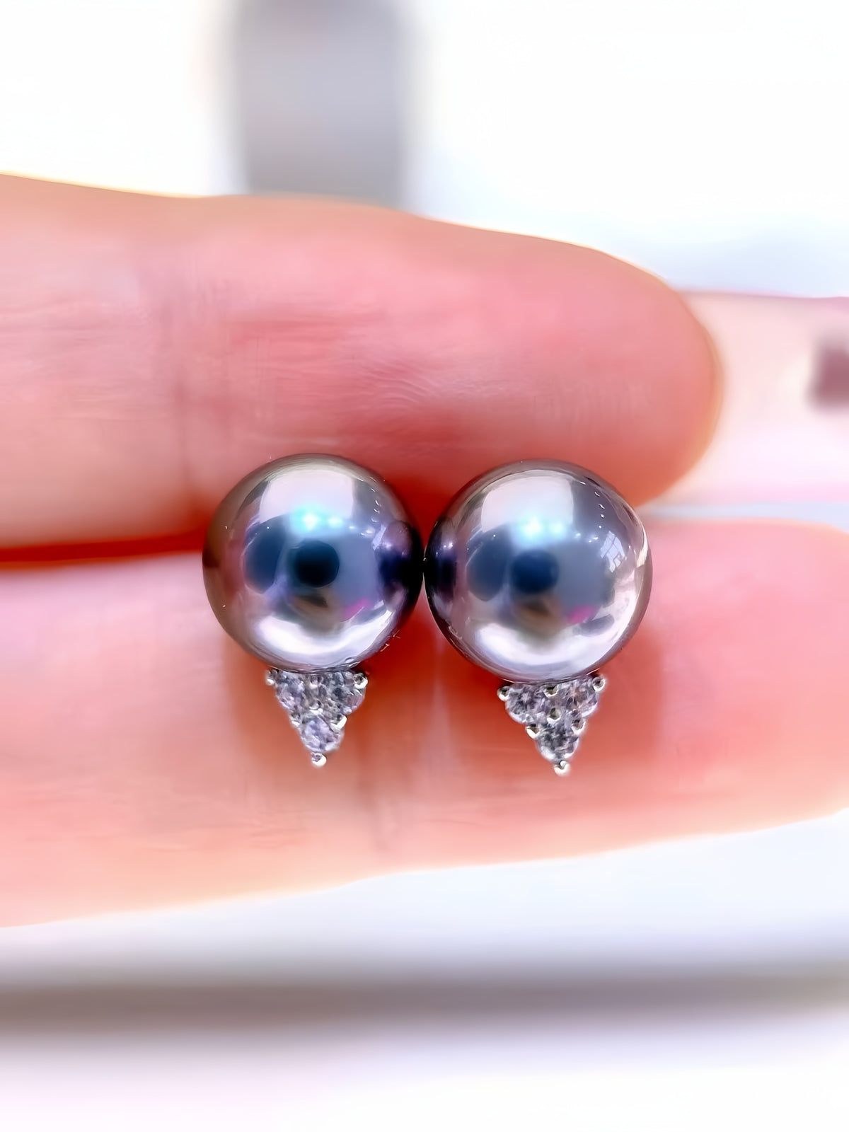 A pair of stud earrings with round dark gray pearl-like beads and small silver triangular crystal clusters, shown resting on a person's fingers against a bright white background.