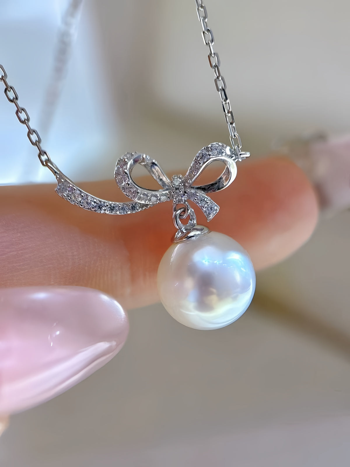 A close-up of a silver necklace with a bow-shaped pendant set with small clear stones and a dangling white pearl, held between fingers.