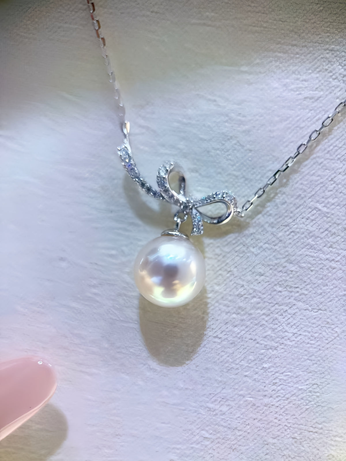 A silver pendant necklace featuring a bow-shaped setting with small clear stones and a single lustrous pearl pendant, displayed on a textured light surface.
