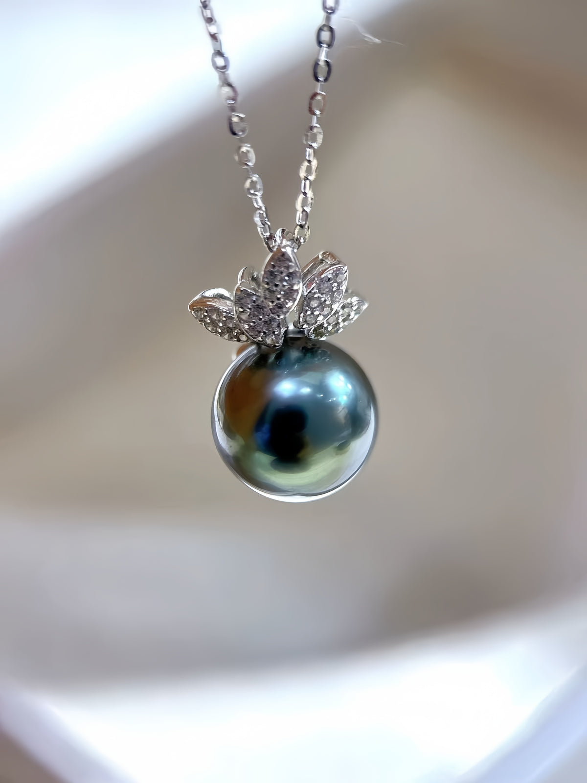 A close-up of a Tasaki pearl necklace featuring a dark blue-gray round pearl pendant set in a silver butterfly-shaped halo with small clear stones, hanging against a white background.
