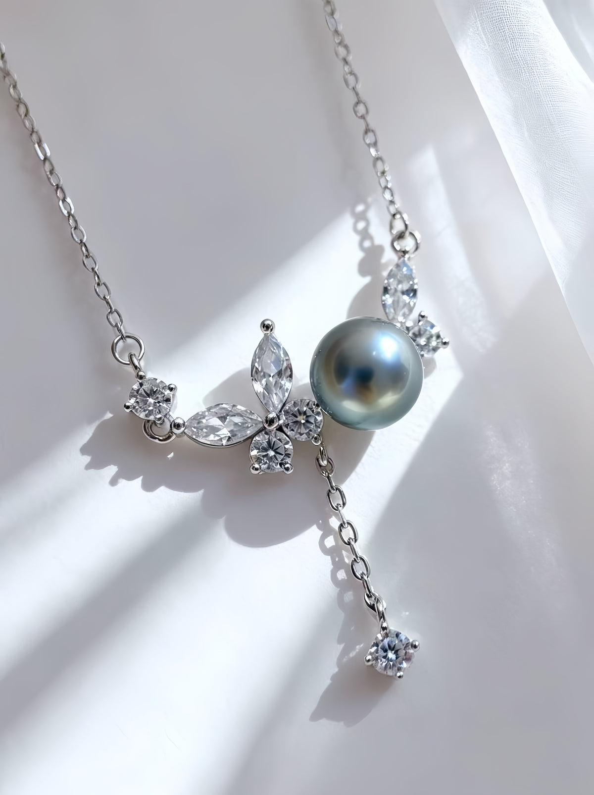 A silver-toned necklace featuring a large round gray pearl and clear crystal floral and butterfly motifs, displayed on a white draped fabric background with soft shadows.