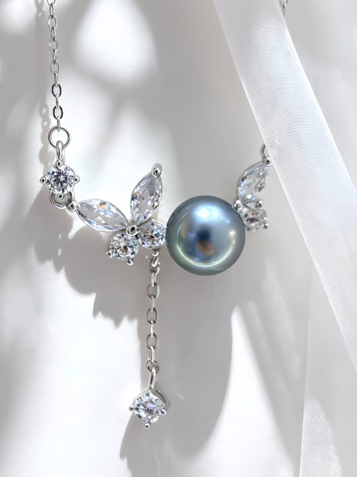 A silver-toned necklace featuring a large round gray-blue pearl at the center, flanked by butterfly-shaped crystal clusters and a dangling clear crystal charm, displayed on a white draped fabric background.