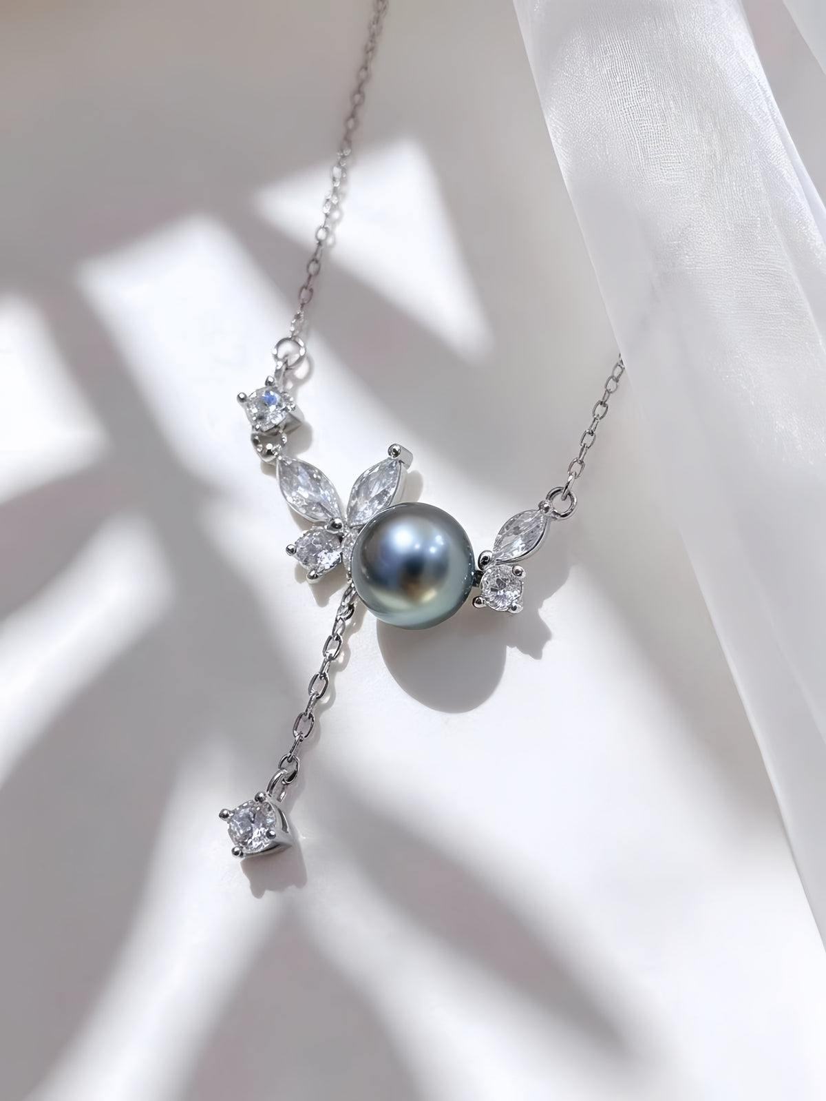 A silver-toned necklace featuring a central dark gray pearl pendant, surrounded by clear marquise and round crystals, with a dangling clear crystal charm, displayed on a white fabric background with soft leaf shadows.
