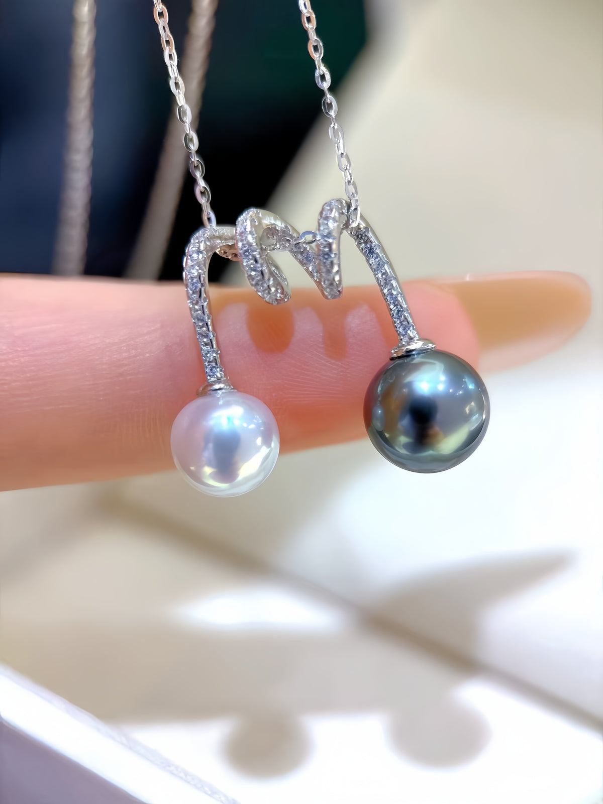 A close-up of a silver pendant necklace held on a finger. The necklace features a curved silver chain with small clear stones and two pearl drops: one white and one dark gray/black Tahitian pearl.