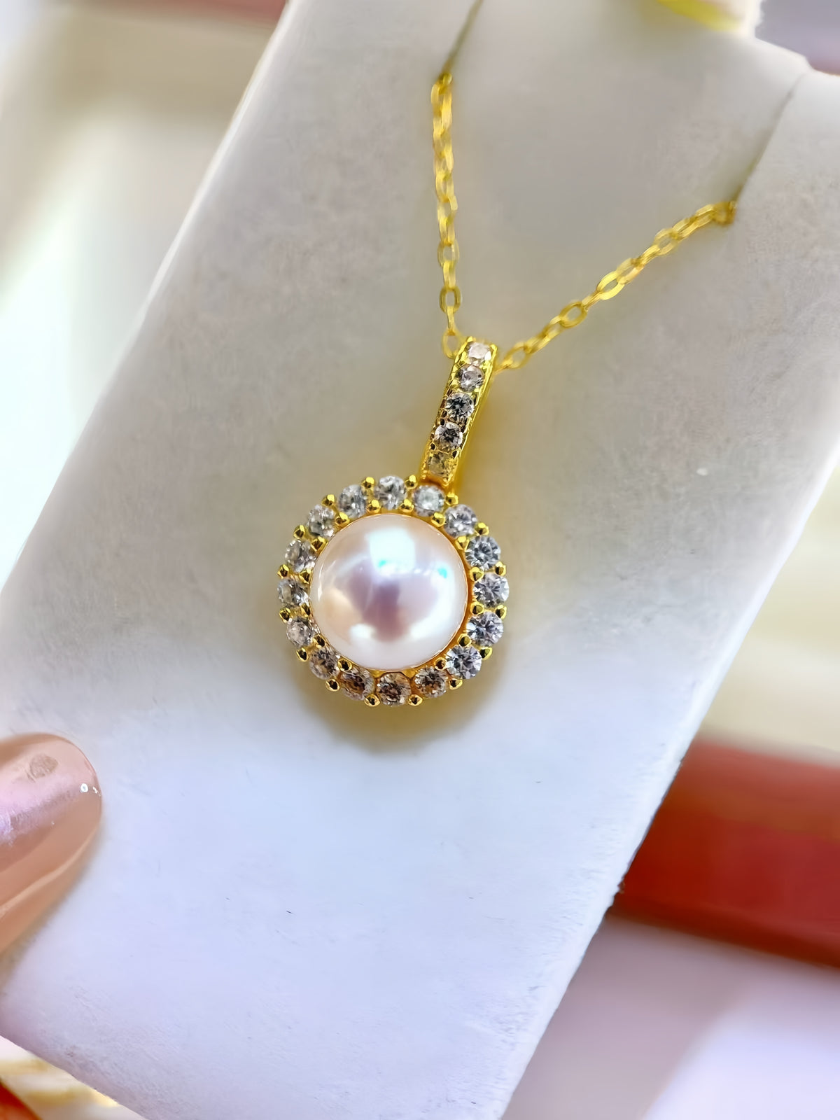 A gold pendant necklace with a central iridescent pearl surrounded by small clear stones, held between fingers against a soft white background.