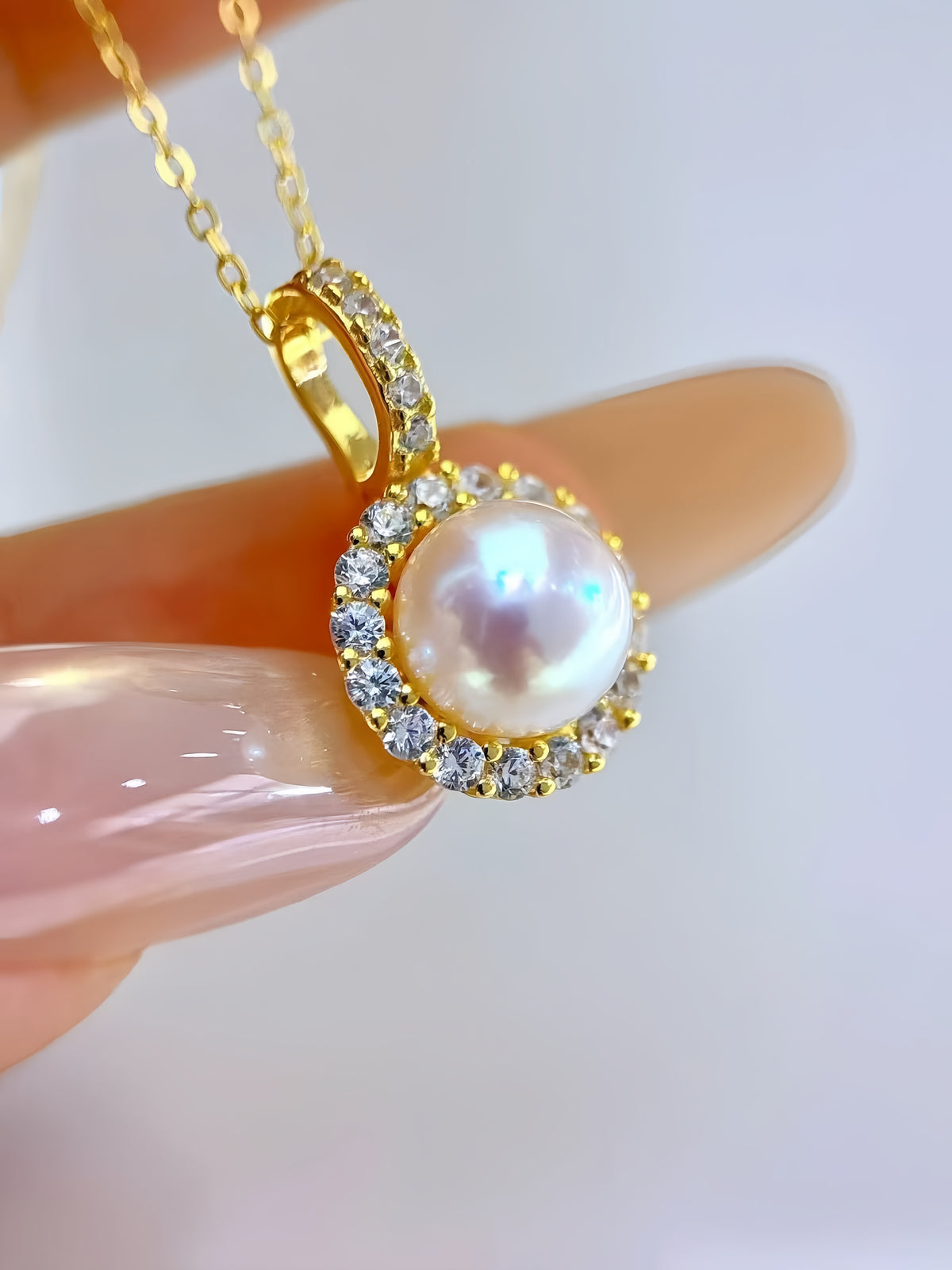 A gold pendant necklace with a central iridescent pearl surrounded by small clear stones, held between fingers against a soft white background.
