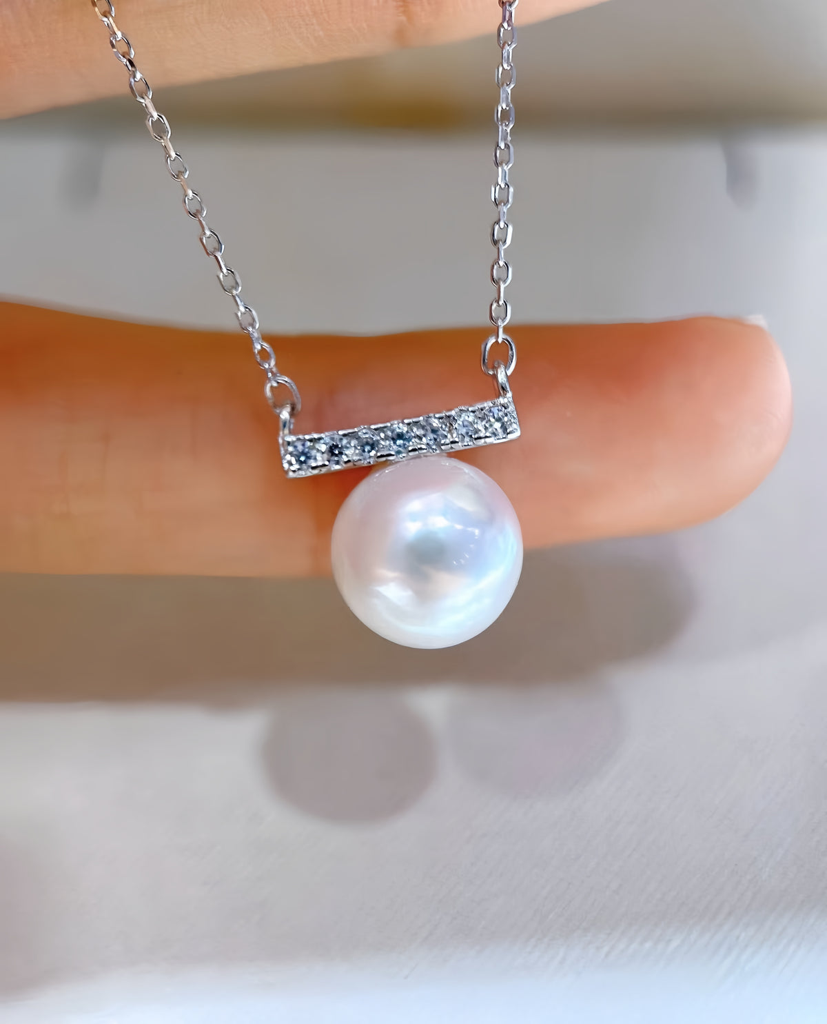 A close-up of a delicate silver necklace with a bar-set of small clear stones and a single lustrous white pearl pendant, held against a soft light background.