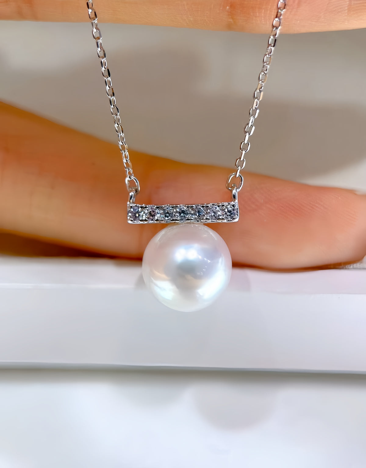 A silver bar necklace with a single large white pearl pendant, held against a soft-focus background of skin and a white surface.