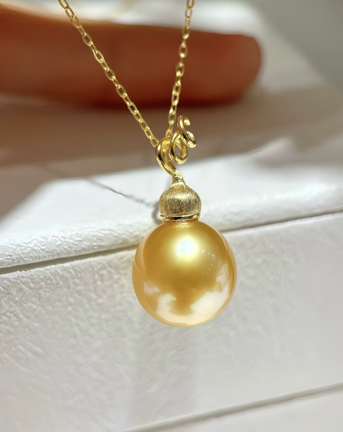 A gold pendant necklace featuring a large, lustrous golden pearl suspended from a delicate gold chain, resting on a white textured surface with a soft-focus background.