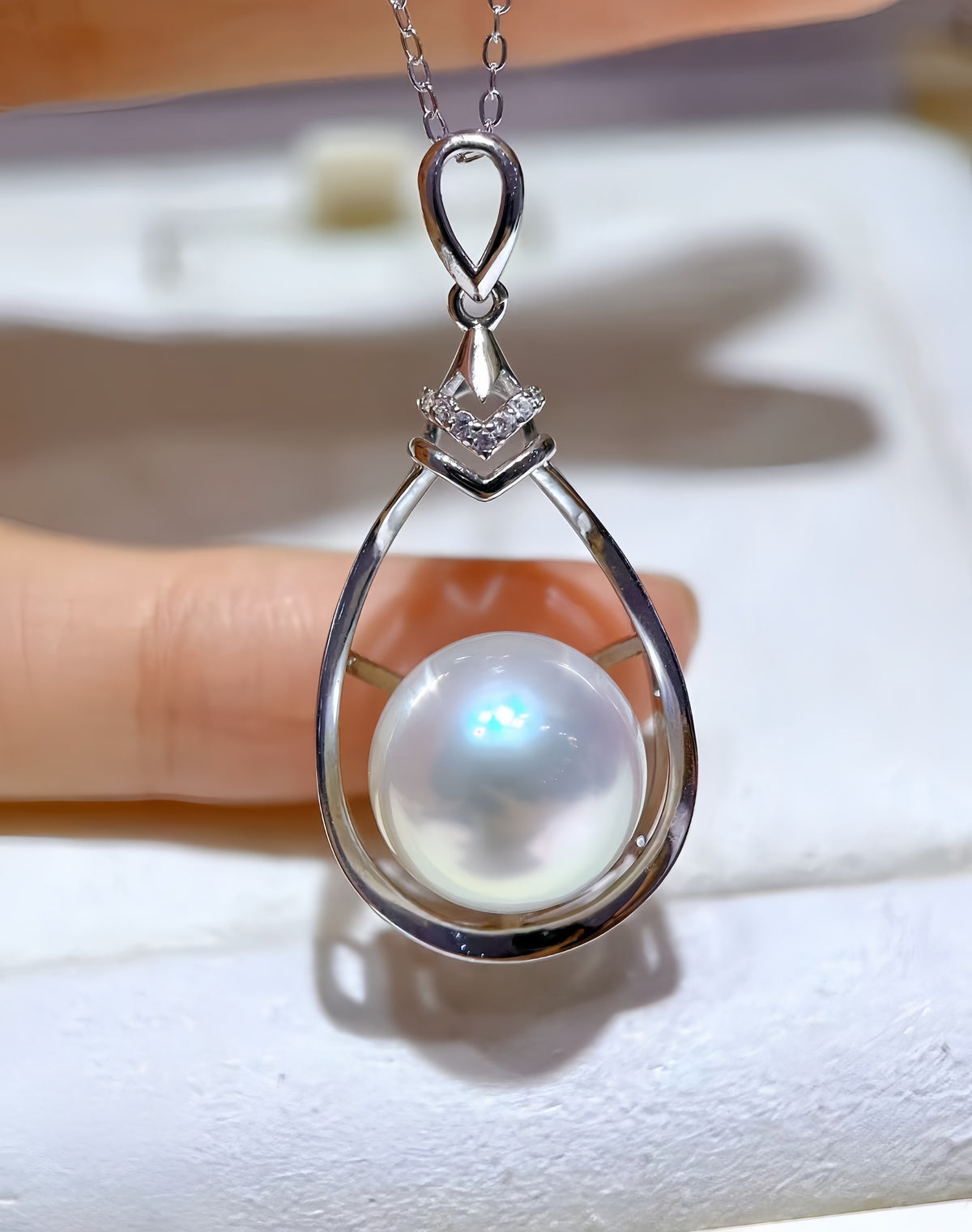A teardrop-shaped pearl pendant in a silver-toned setting, displayed on a white jewelry card inside an open cream-colored presentation box.