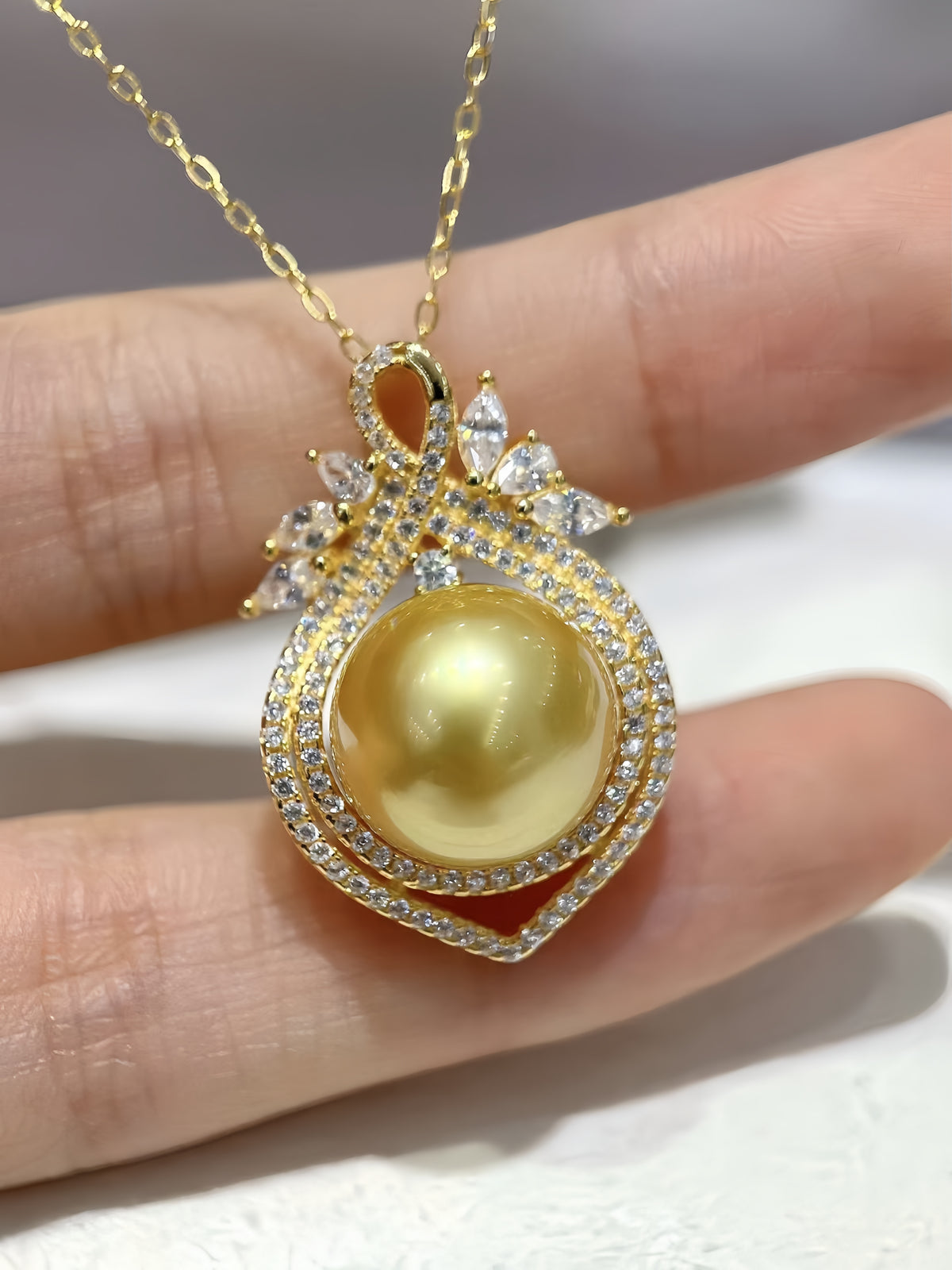 A gold pendant necklace with a large round yellow pearl in a diamond-encrusted teardrop setting, held on a person's fingers against a light background.