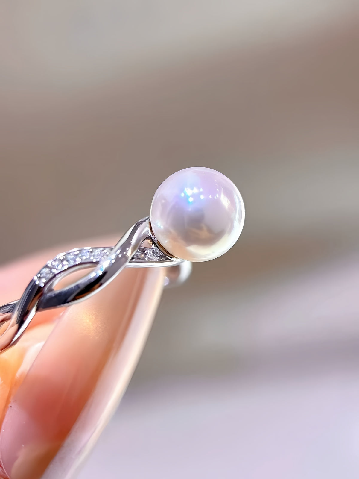 A close-up of a silver ring with a twisted band and a single round white pearl, held between fingers against a soft beige background.
