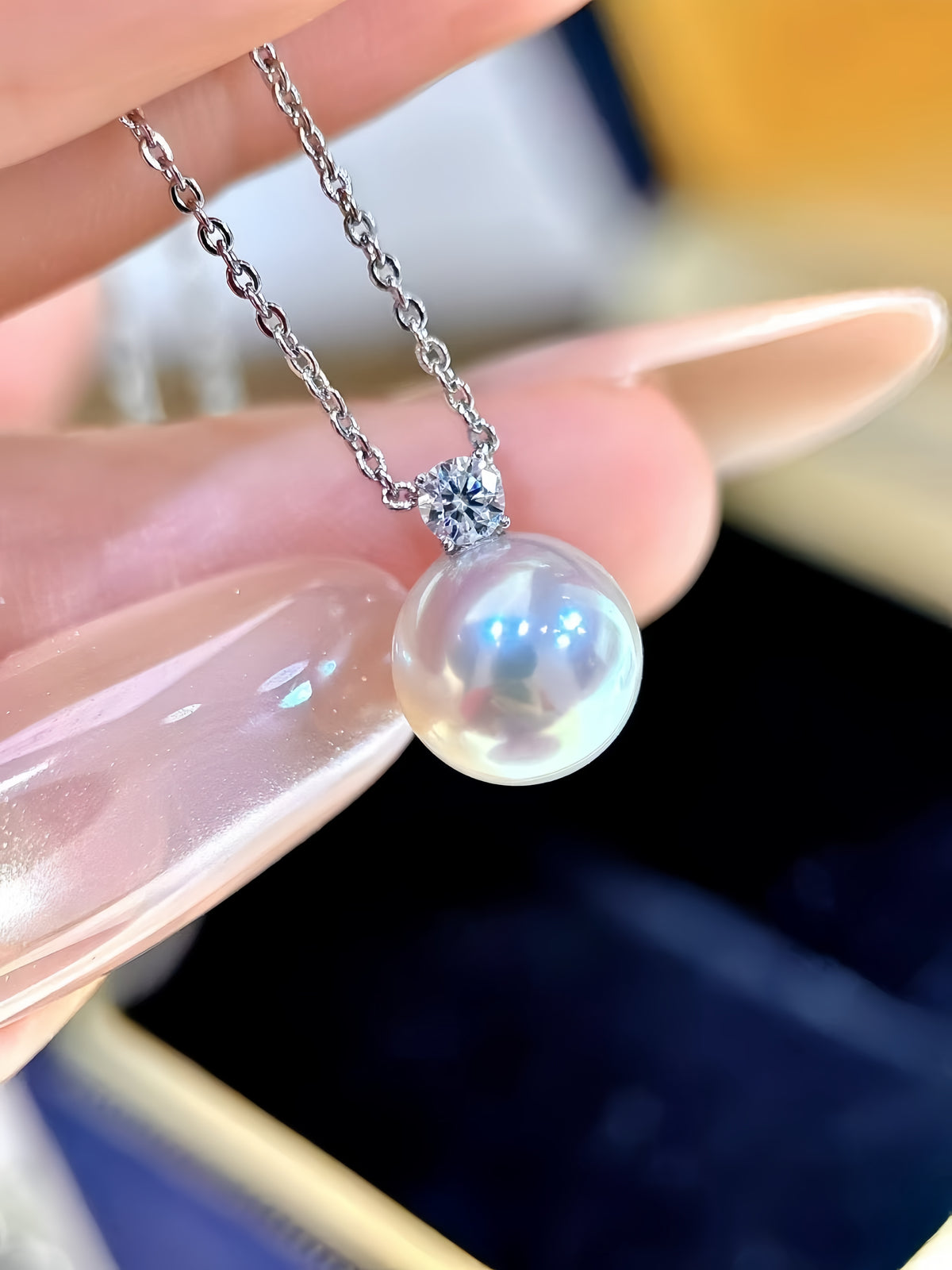 A close-up of a pearl pendant necklace with a silver-toned chain and bezel setting, held between fingers against a softly blurred background.