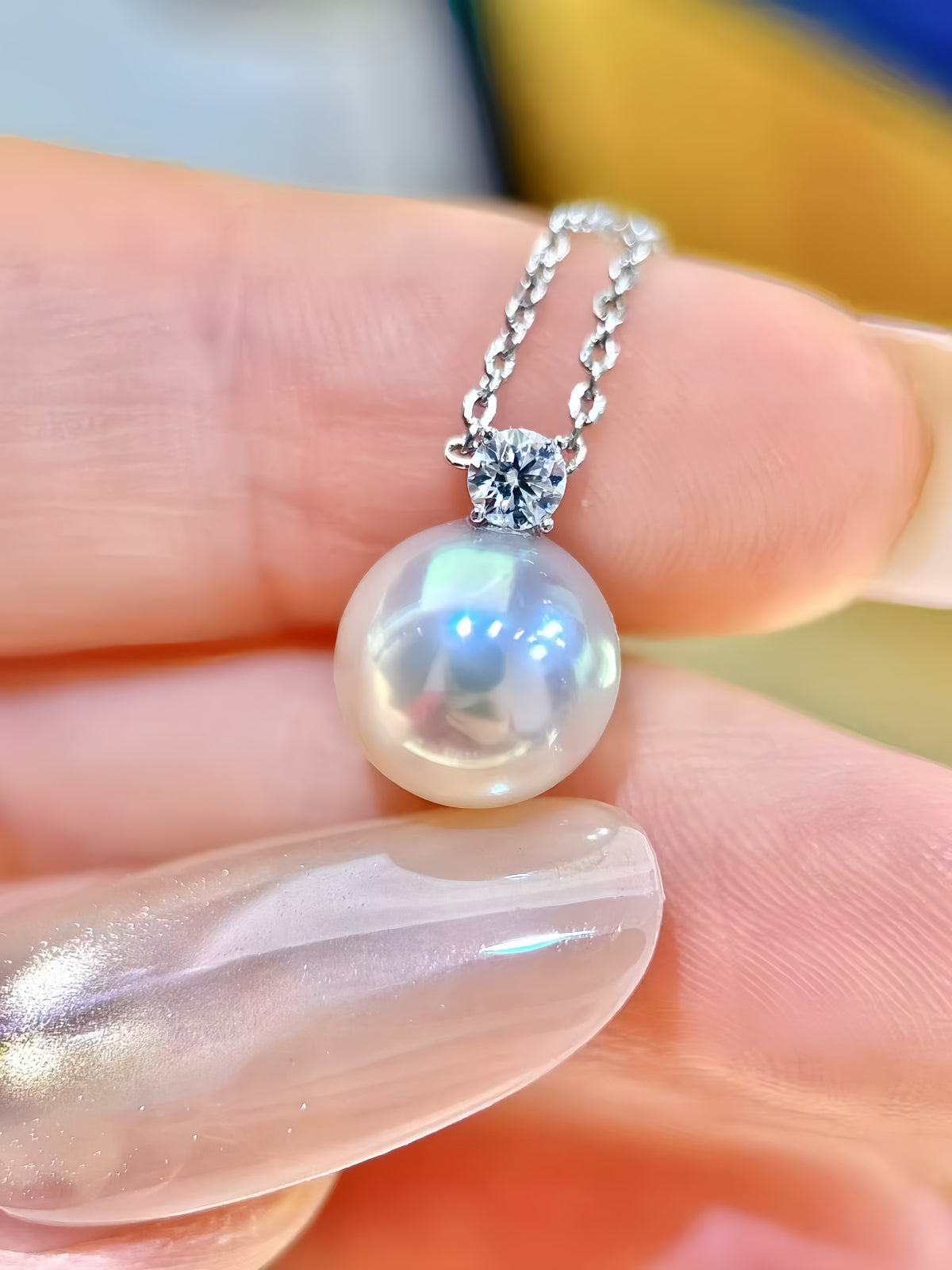 A close-up of a pearl pendant necklace with a silver-toned chain and bezel setting, held between fingers against a softly blurred background.