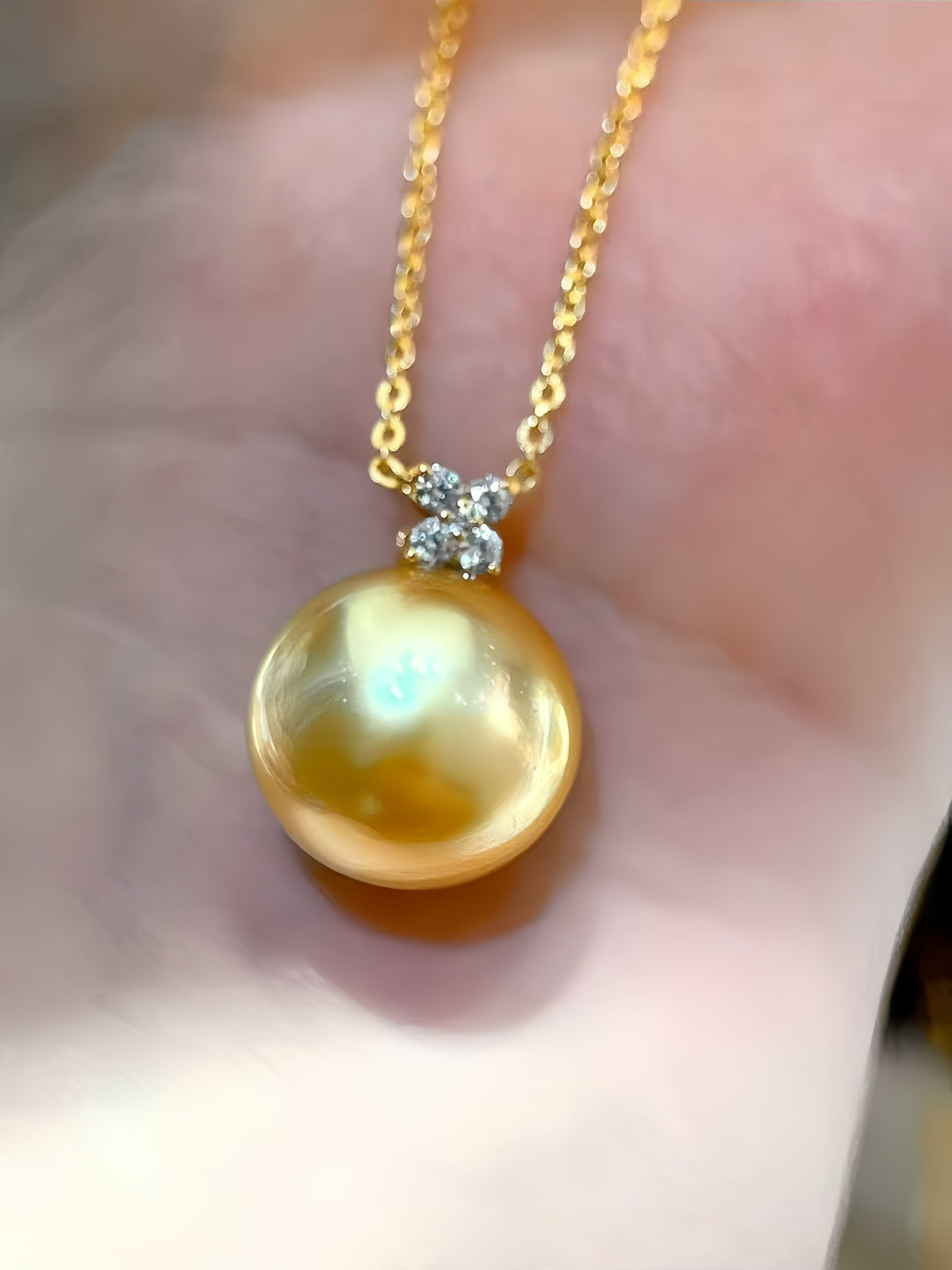 A close-up of a gold chain necklace with a large, round, lustrous gold pearl pendant, held against a soft-focus skin background.
