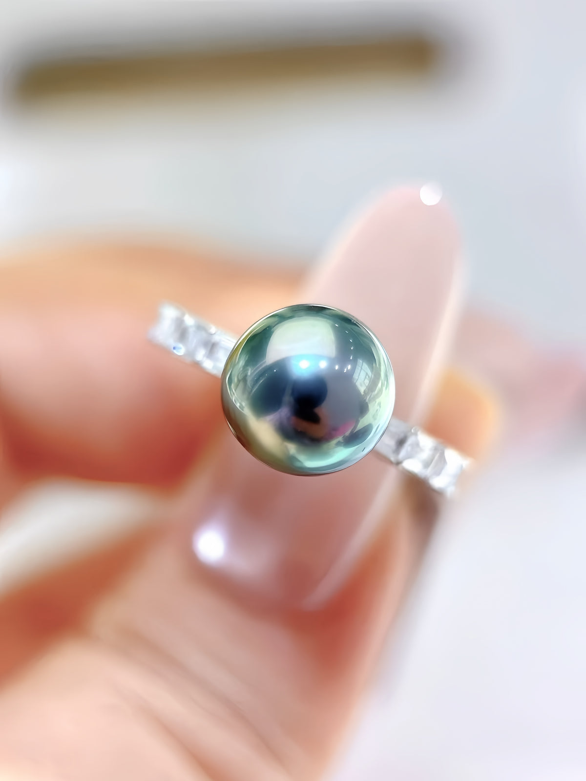 A close-up of a silver ring featuring a round, lustrous greenish-blue pearl, held between fingers against a soft, blurred light background.