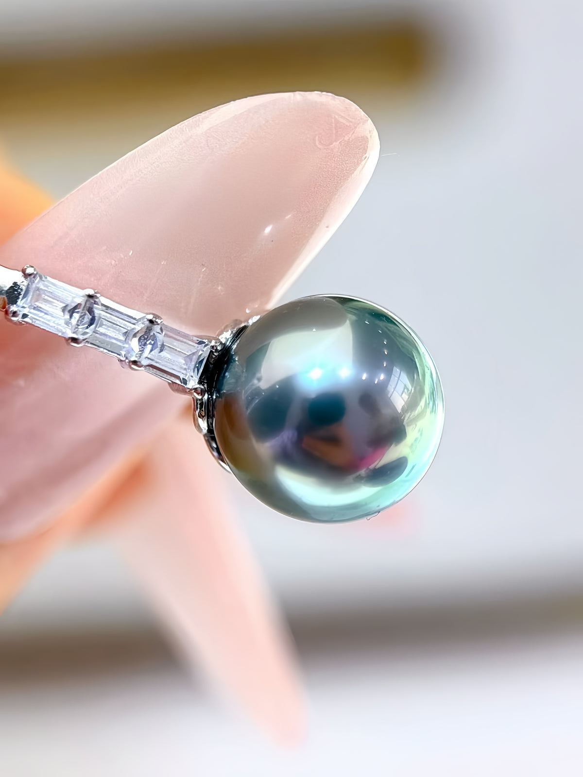 A close-up of a silver ring with a round, iridescent green-blue pearl and clear baguette side stones, held between fingers.