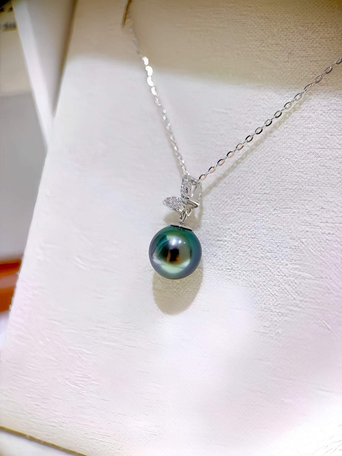 A silver necklace with a single round dark green pearl pendant and a small clear stone accent, displayed on a white jewelry bust.