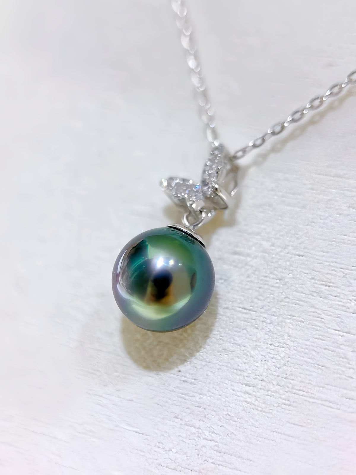 A silver pendant necklace featuring a round, iridescent green-black Tahitian pearl and a silver butterfly-shaped setting with small clear stones, shown on a textured white surface.