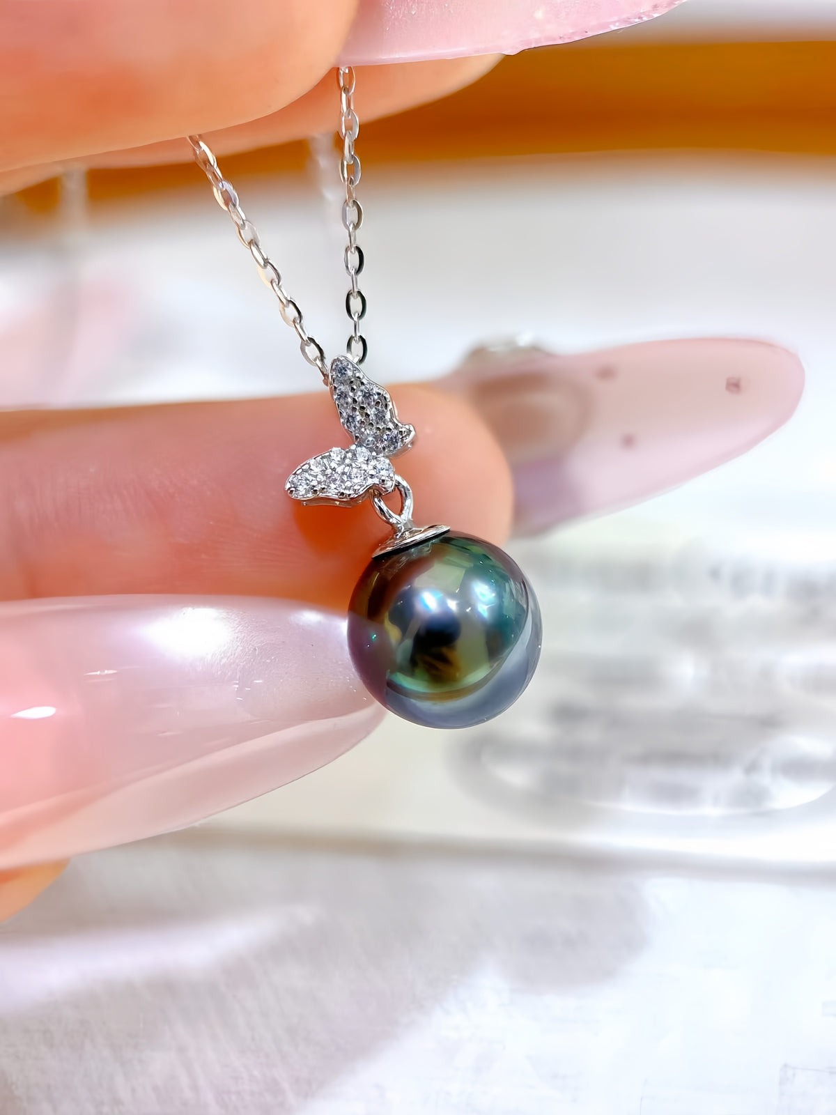 A close-up of a hand holding a silver pendant necklace with a large, iridescent dark green-black freshwater pearl and a small crystal-embellished silver leaf charm.