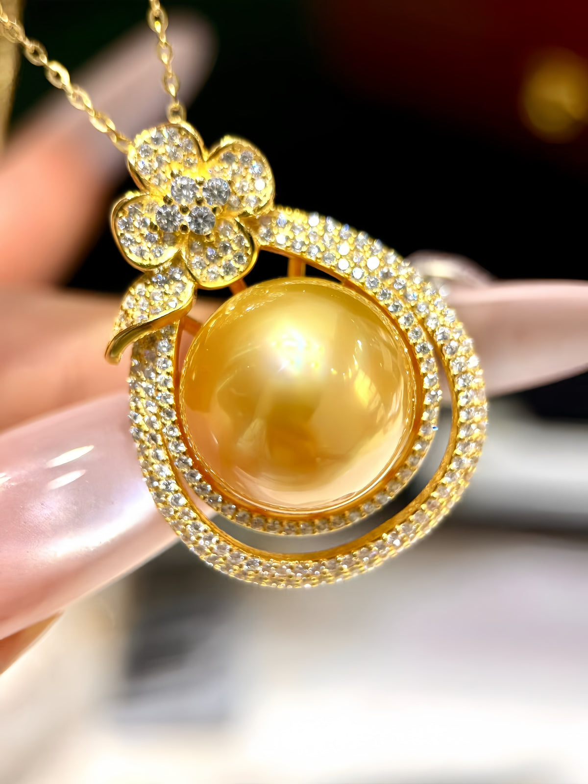 A gold pendant necklace featuring a large round yellow-gold pearl surrounded by pavé-set clear stones, held by a hand. The pendant includes a flower-shaped top set with small clear stones.