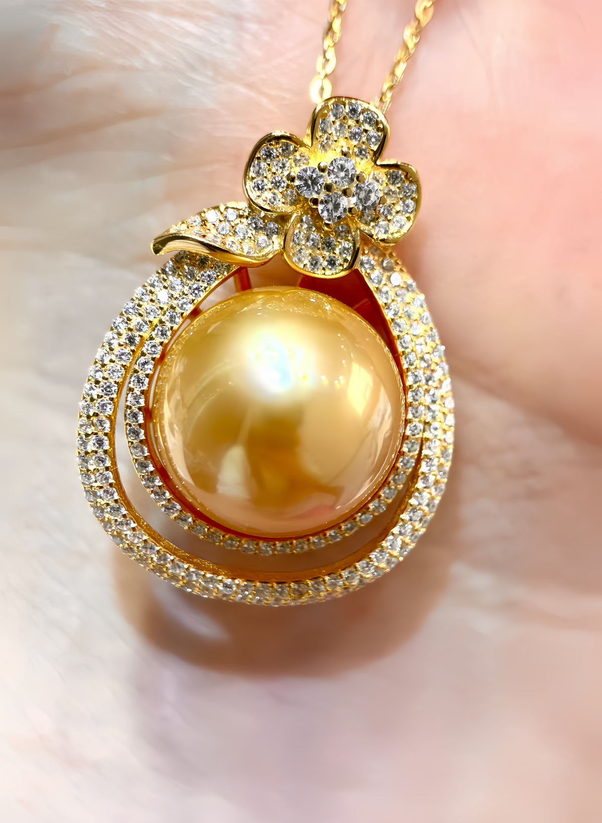 A gold pendant necklace featuring a large round golden pearl surrounded by double rows of pavé-set clear stones, with a flower-shaped top accented by small clear stones, shown resting on a soft pink surface.