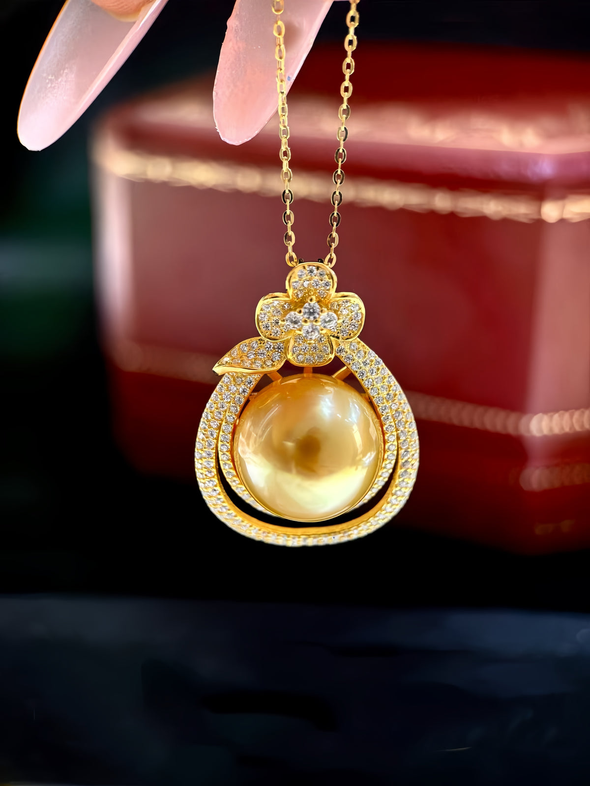 A gold pendant necklace featuring a large round yellow pearl surrounded by a pavé-set gold frame, held by a hand with long pink nails in front of a blurred red jewelry box.
