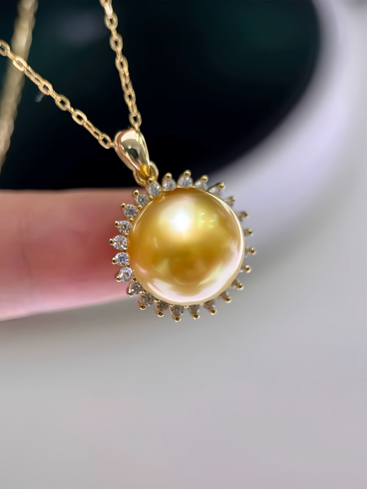 A gold pendant necklace featuring a large, lustrous yellow pearl surrounded by small clear stones, held by a finger against a soft-focus background.