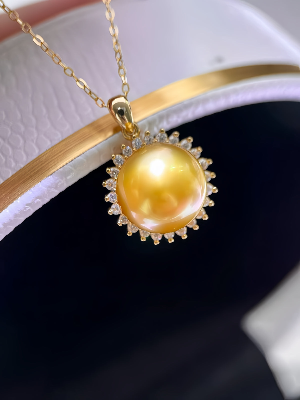 A gold pendant necklace featuring a large, lustrous yellow pearl surrounded by small clear stones, held by a finger against a soft-focus background.