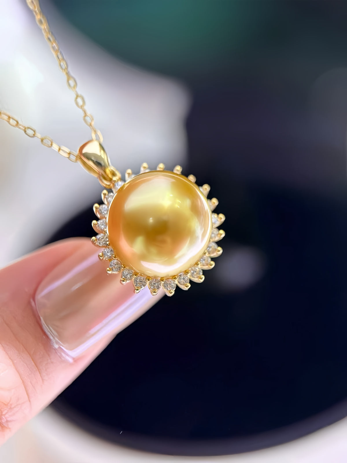 A gold pendant necklace featuring a large, lustrous yellow pearl surrounded by small clear stones, held by a finger against a soft-focus background.