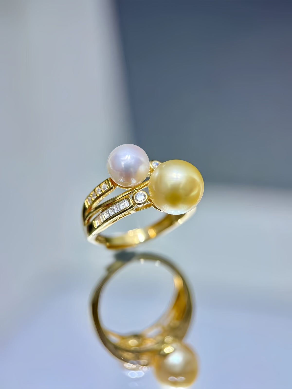 A gold ring featuring two round pearls, one pale pinkish-white and one golden-yellow, accented with small clear stones, shown on a reflective light surface.