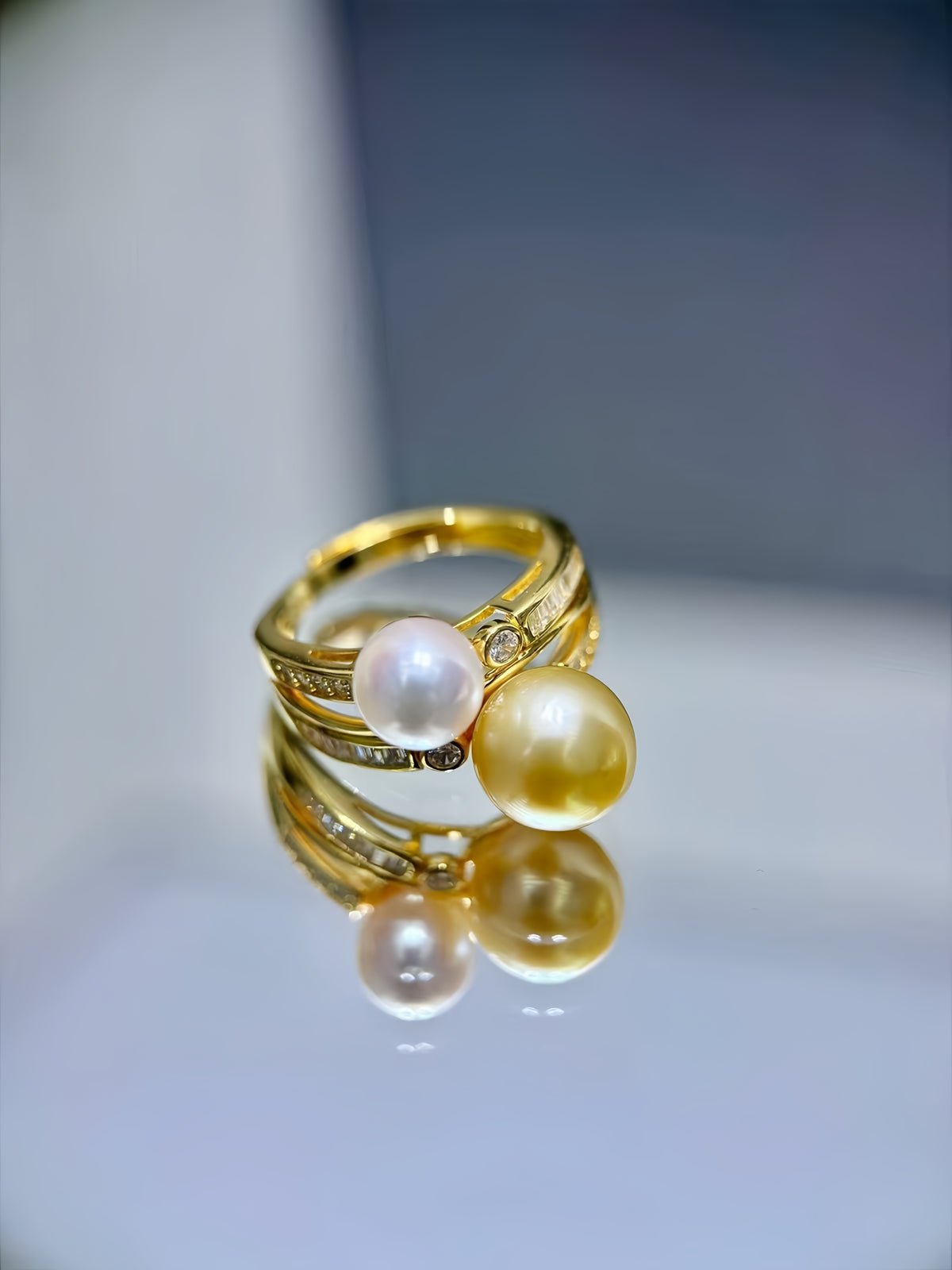 A gold ring featuring two large round pearls, one white and one yellow, with small inset stones along the band, displayed on a reflective surface.
