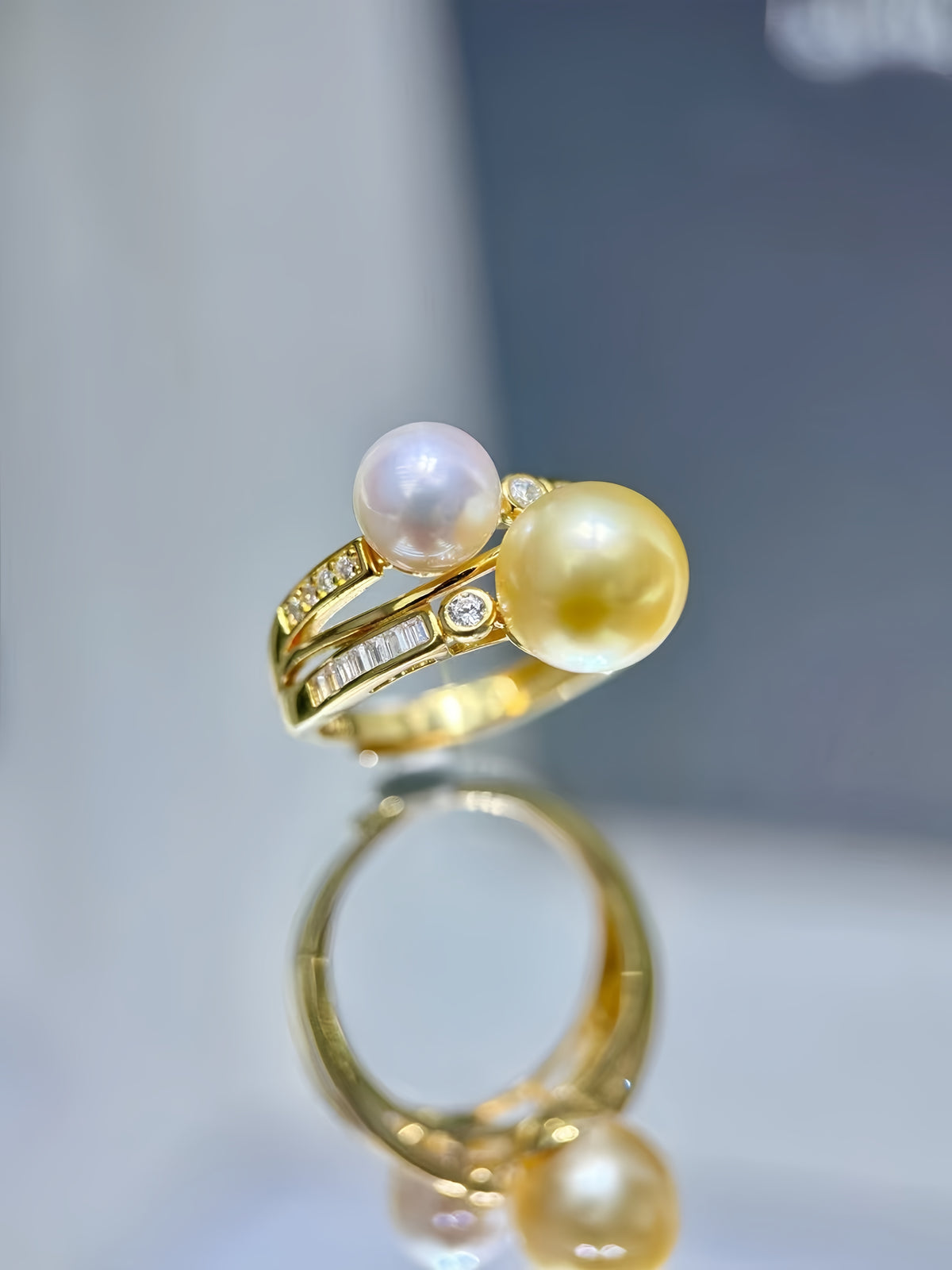 A gold ring featuring two large pearls, one white and one yellow, accented with small clear stones, displayed on a reflective surface.