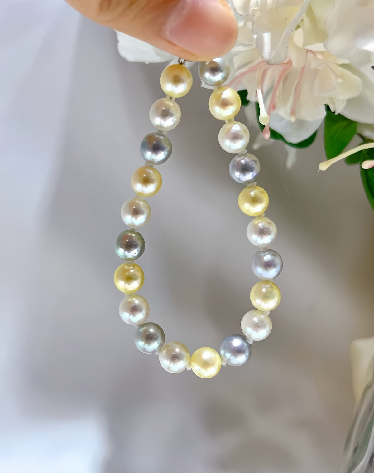 A close-up of a pearl necklace being held by a hand, featuring a mix of white, yellow, and gray pearls, with white flowers in the background.