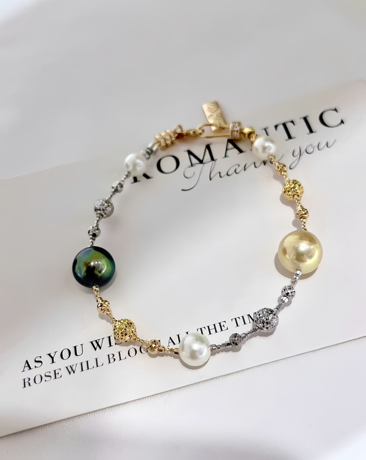 A delicate gold and silver chain bracelet with white pearls, a dark green iridescent bead, and gold heart charms, displayed on a white card with printed text.