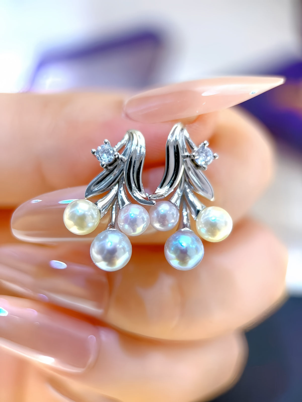 A close-up of a hand with manicured nails holding a pair of silver-toned earrings decorated with iridescent white pearls and small clear stones.