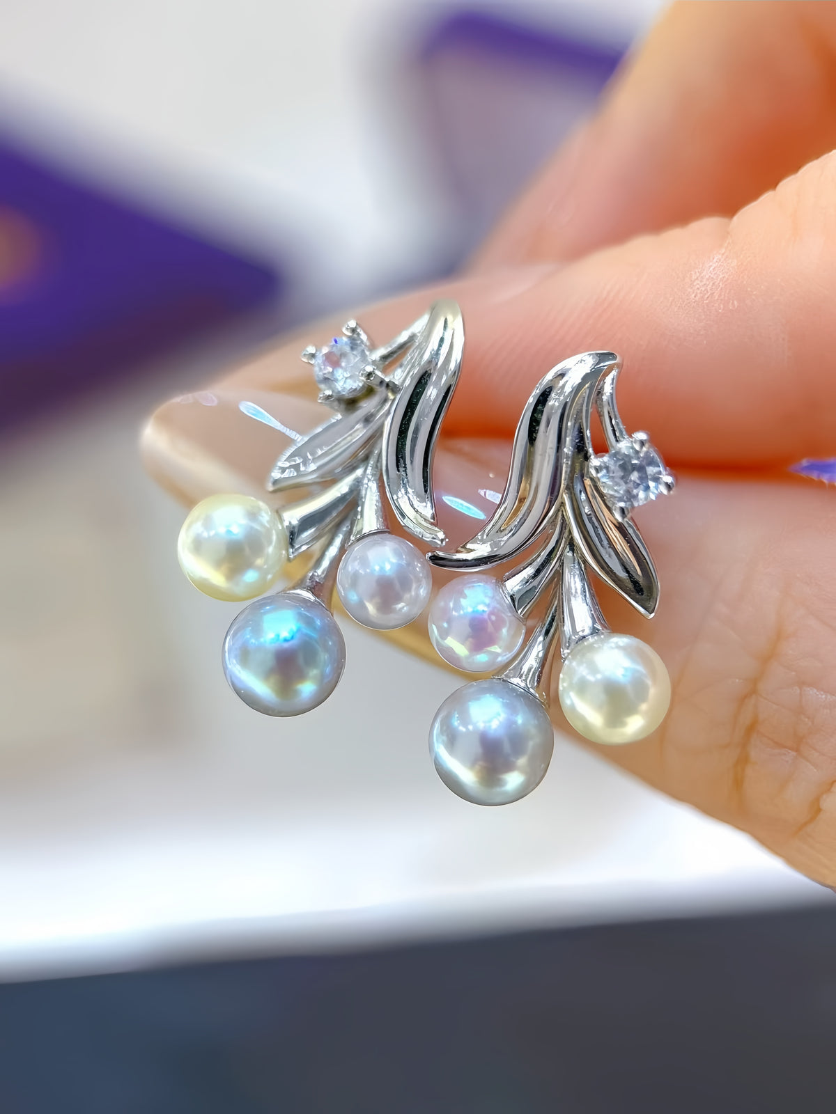 A close-up of a hand holding a pair of silver leaf-shaped stud earrings adorned with iridescent freshwater pearls and small clear stones.