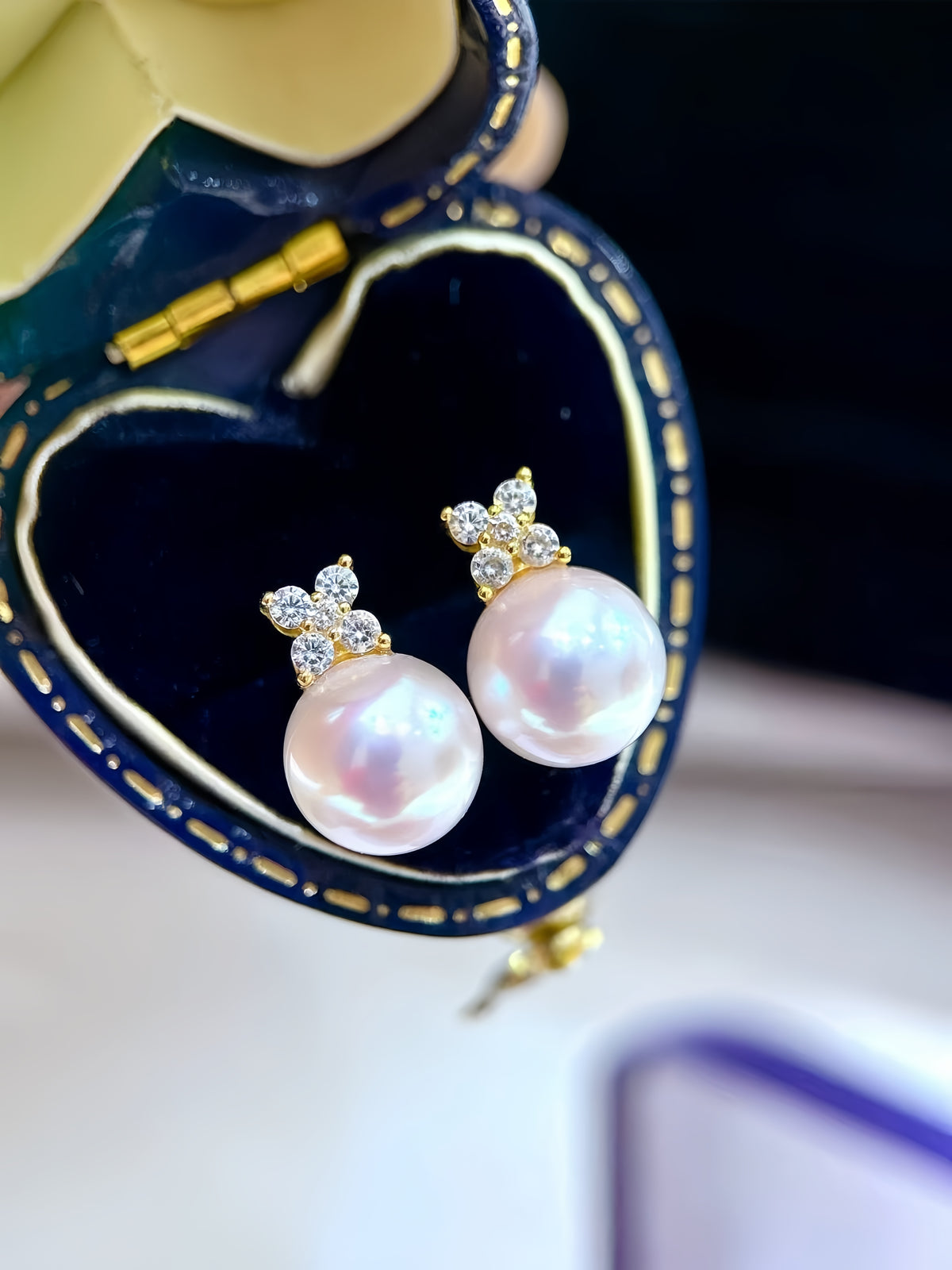 A pair of gold stud earrings with iridescent pink pearl drops and small diamond clusters, displayed inside a heart-shaped navy blue jewelry box.