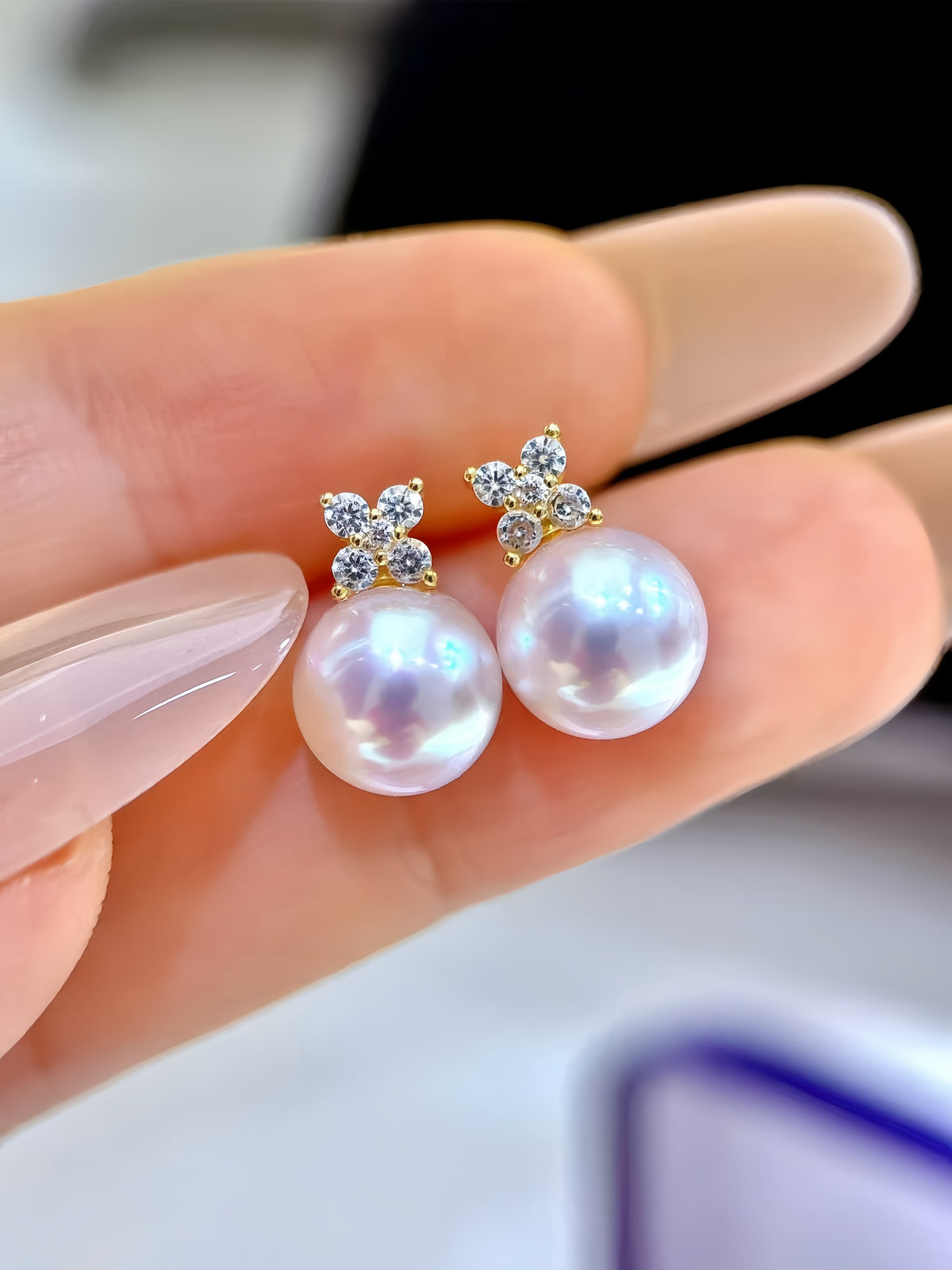 A close-up of a hand holding a pair of gold stud earrings with round white pearls and small clear crystal flower-shaped settings.
