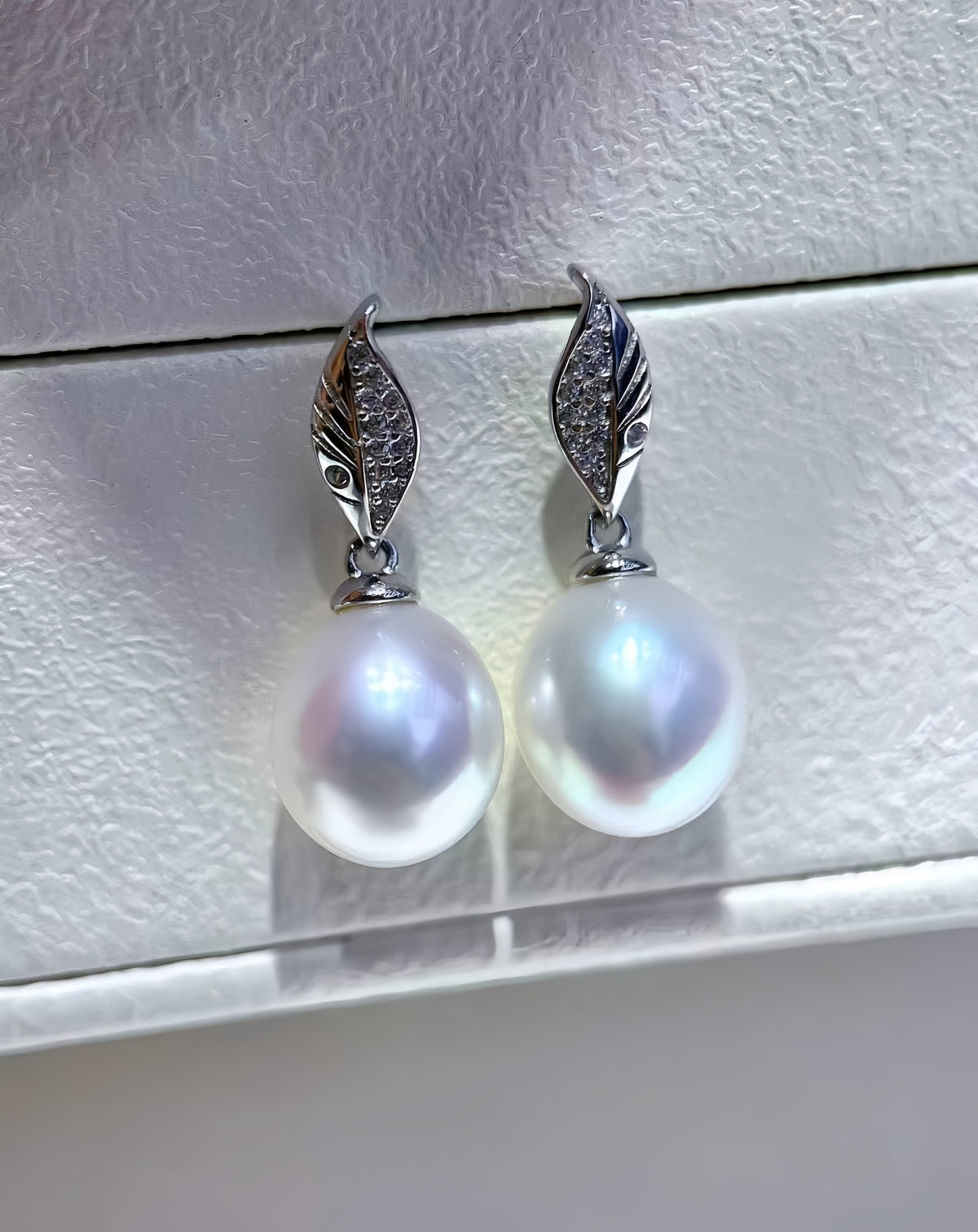 A pair of silver drop earrings featuring round white freshwater pearls and leaf-shaped silver tops set with small clear stones, displayed on a textured light gray surface.