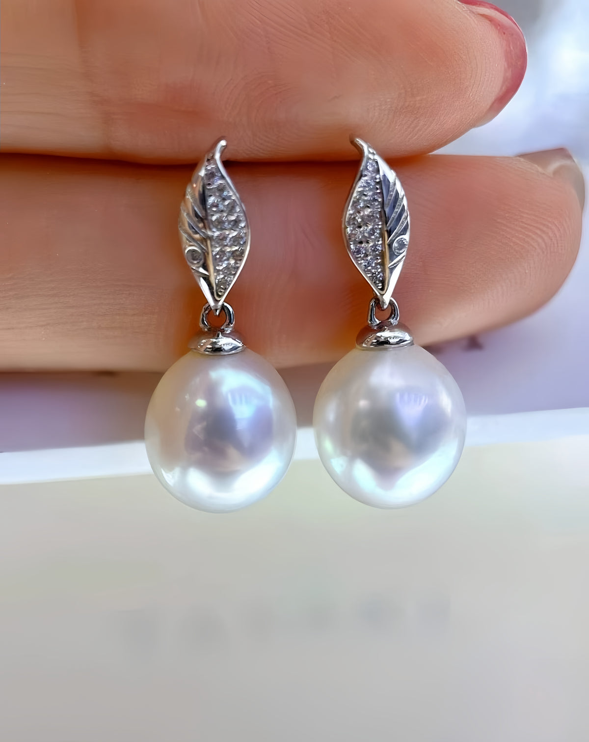 A pair of silver leaf drop earrings with large white freshwater pearls, held between fingers against a light background.