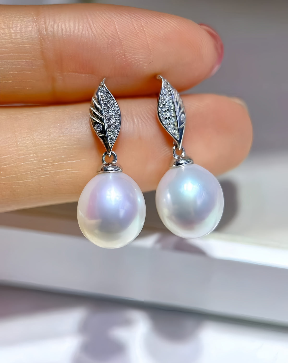 A pair of silver drop earrings with large iridescent white pearls and leaf-shaped crystal-studded tops, held between fingers against a soft light gray background.