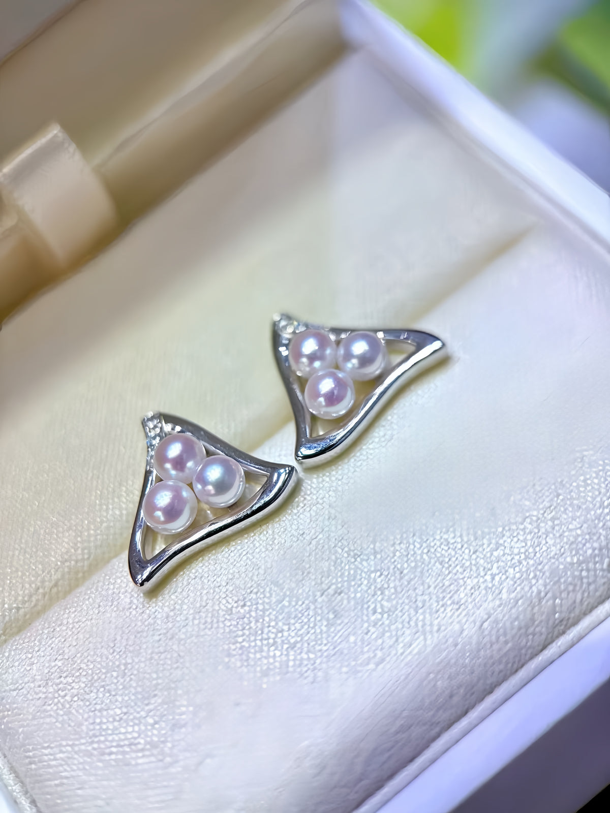 A pair of silver triangle stud earrings with three iridescent white pearls, displayed inside a cream-colored jewelry box.