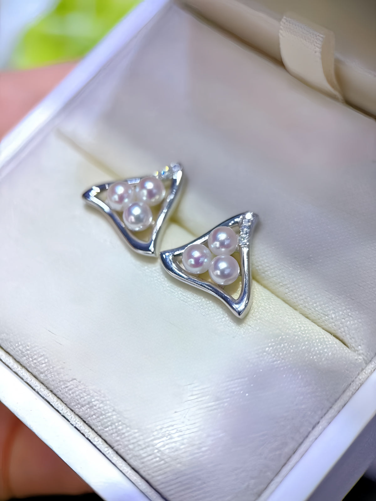 A pair of silver triangular stud earrings with three white pearls and small clear stones, displayed in a cream-colored jewelry box.
