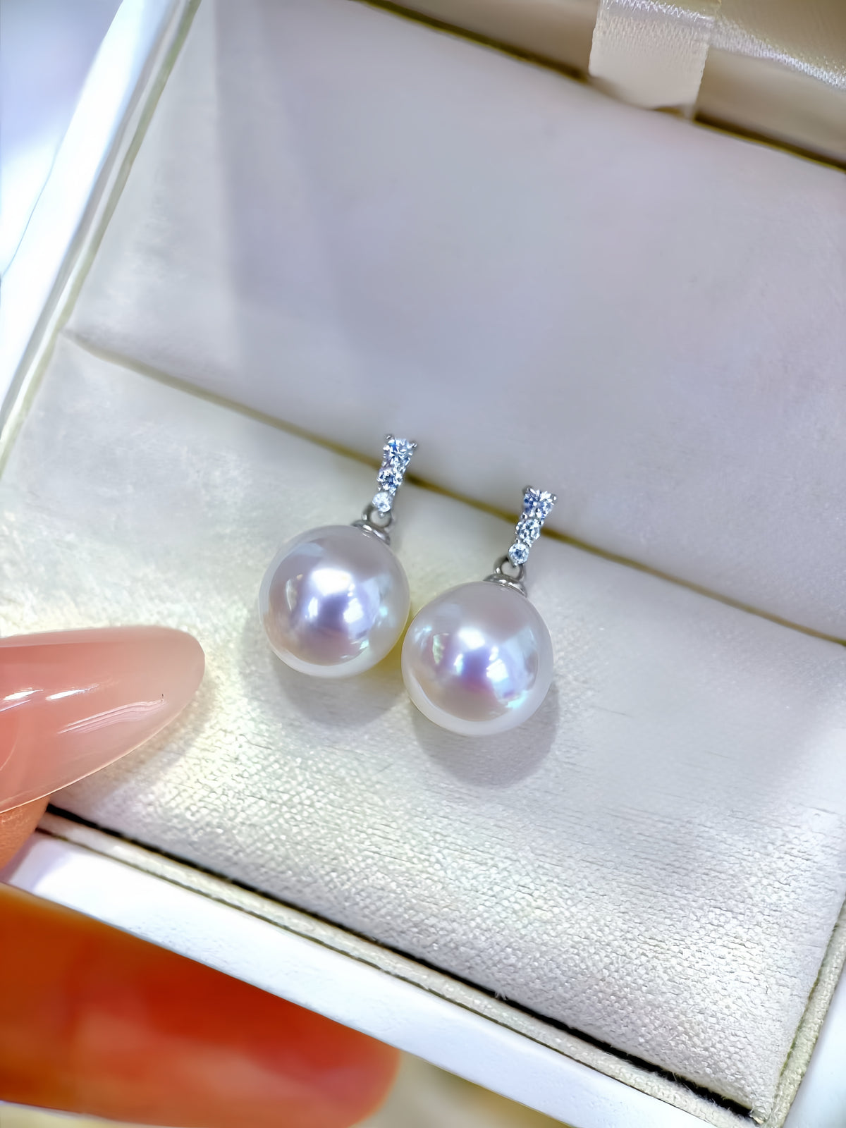 A pair of white freshwater pearl drop earrings with silver-toned pavé hangers, displayed inside a cream-colored jewelry box. A finger with pink manicured nails is visible on the left side of the frame.