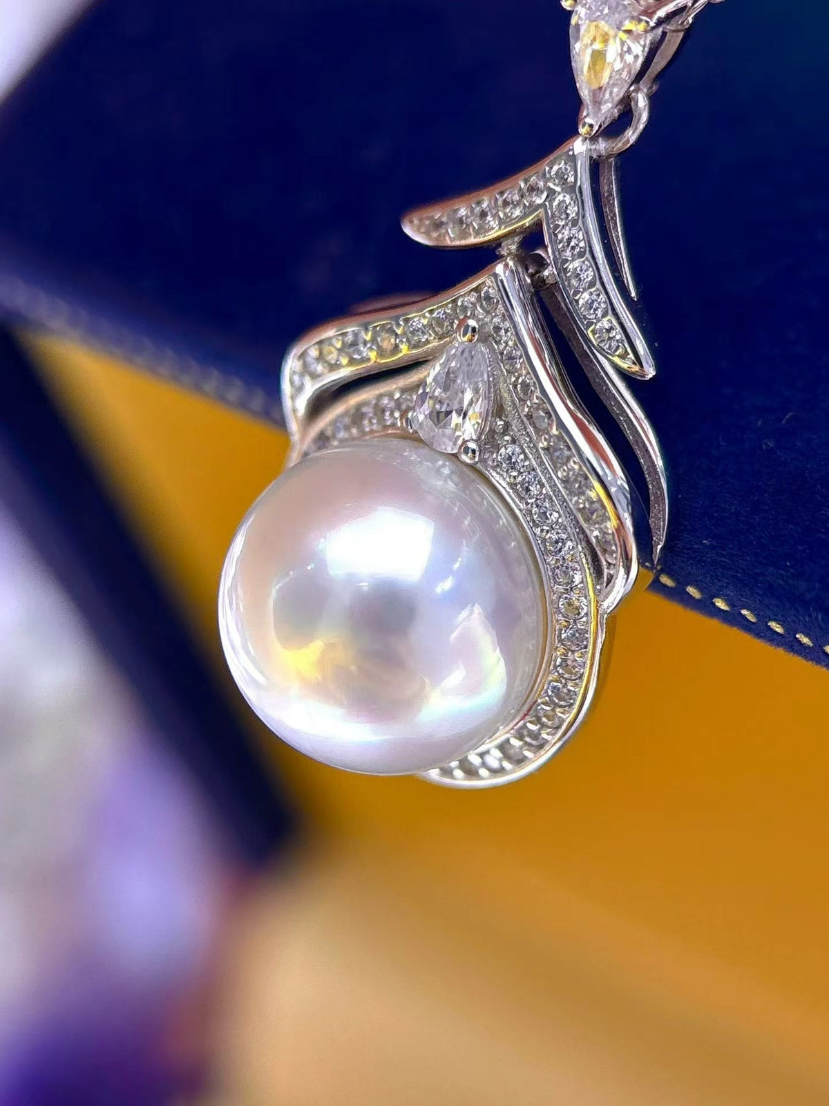A close-up of a silver pendant necklace featuring a large, lustrous white pearl surrounded by small clear stones, resting on a dark blue velvet jewelry box.