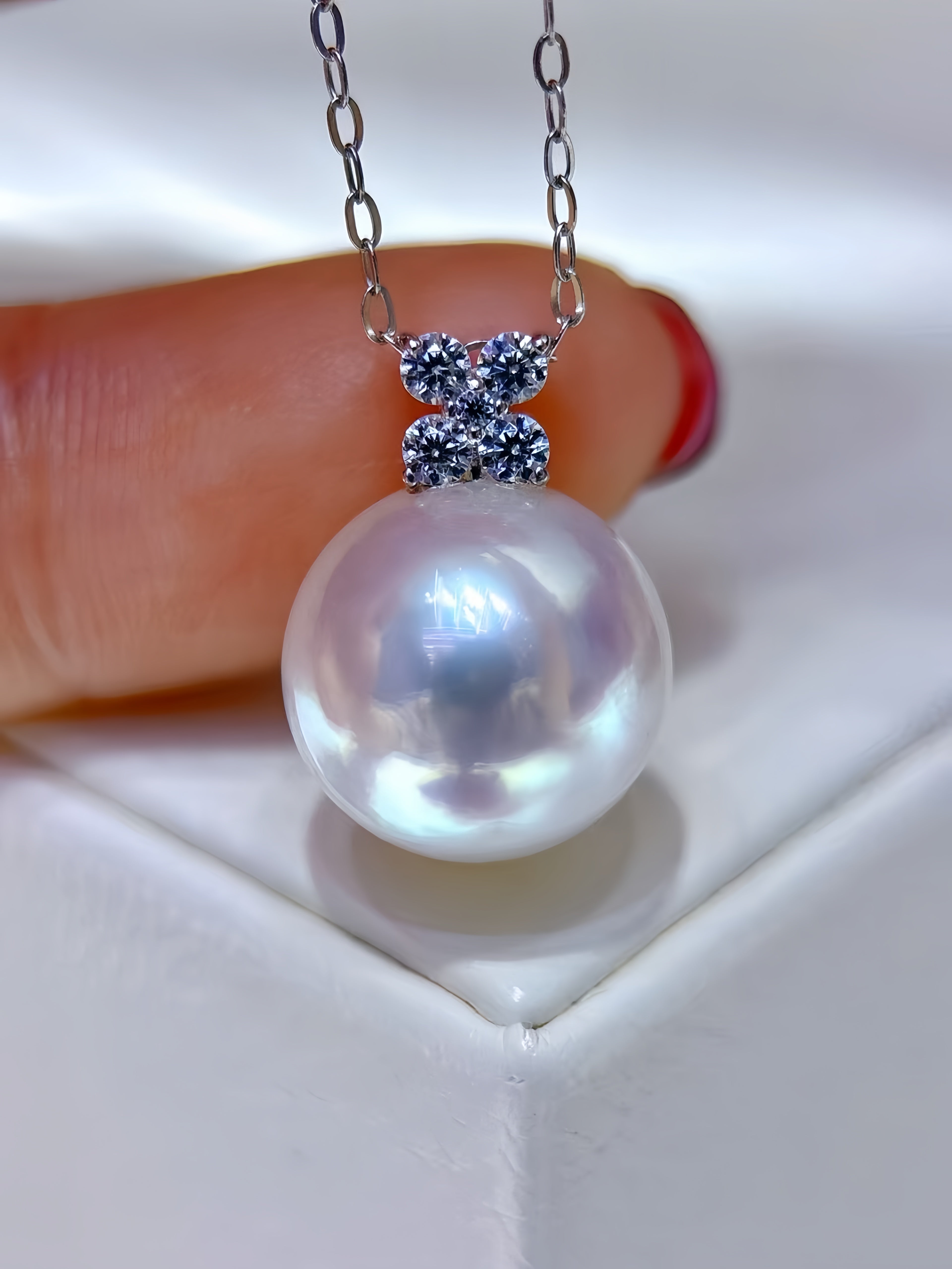 A close-up of a pearl pendant necklace held by a finger. The pendant features a large, lustrous white pearl suspended from a silver chain, with a small silver bow-shaped setting adorned with tiny blue stones.