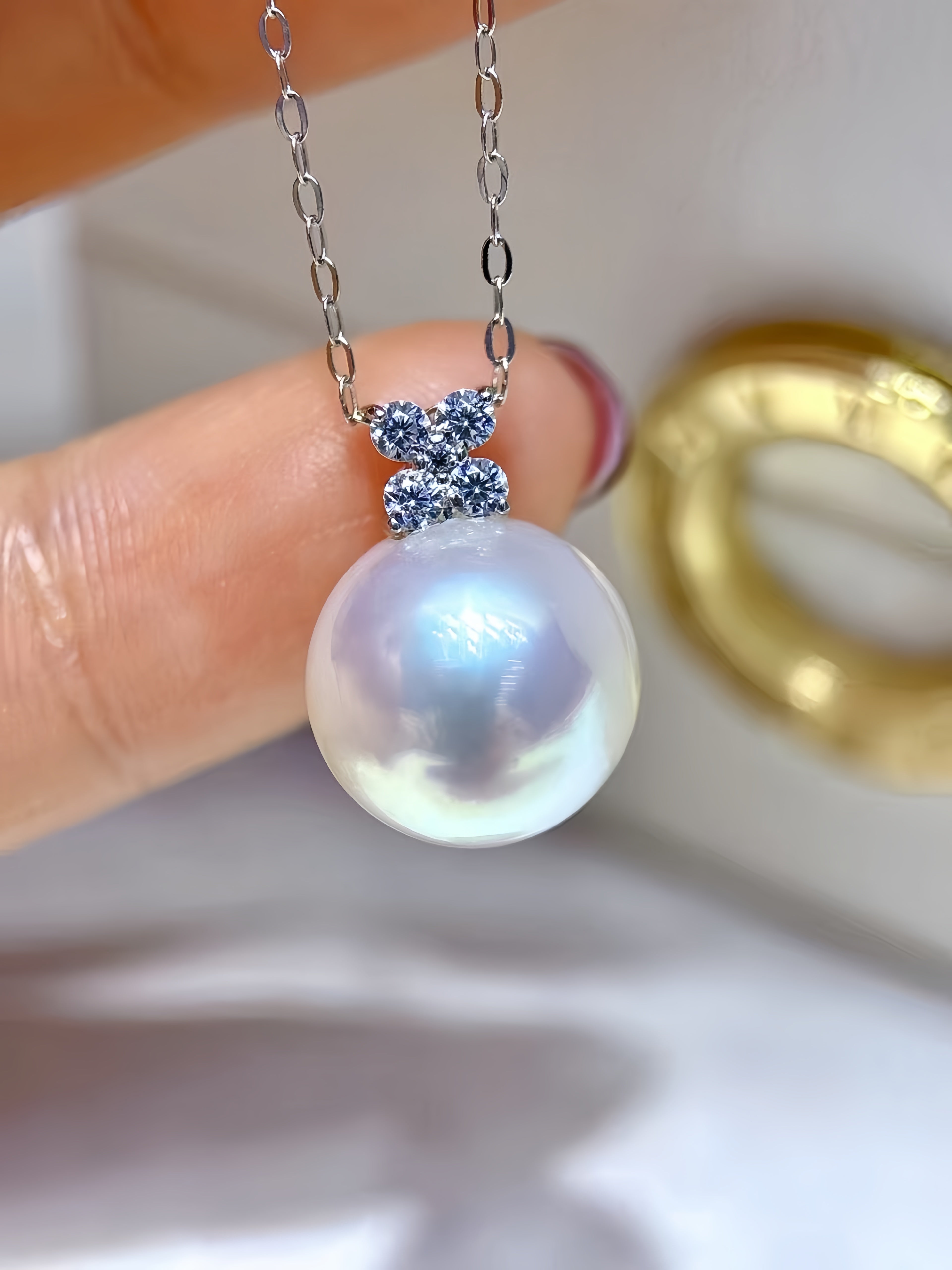 A close-up of a silver chain necklace with a large, lustrous white pearl pendant and a small silver bow-shaped setting accented with sparkling blue stones, held by a hand.