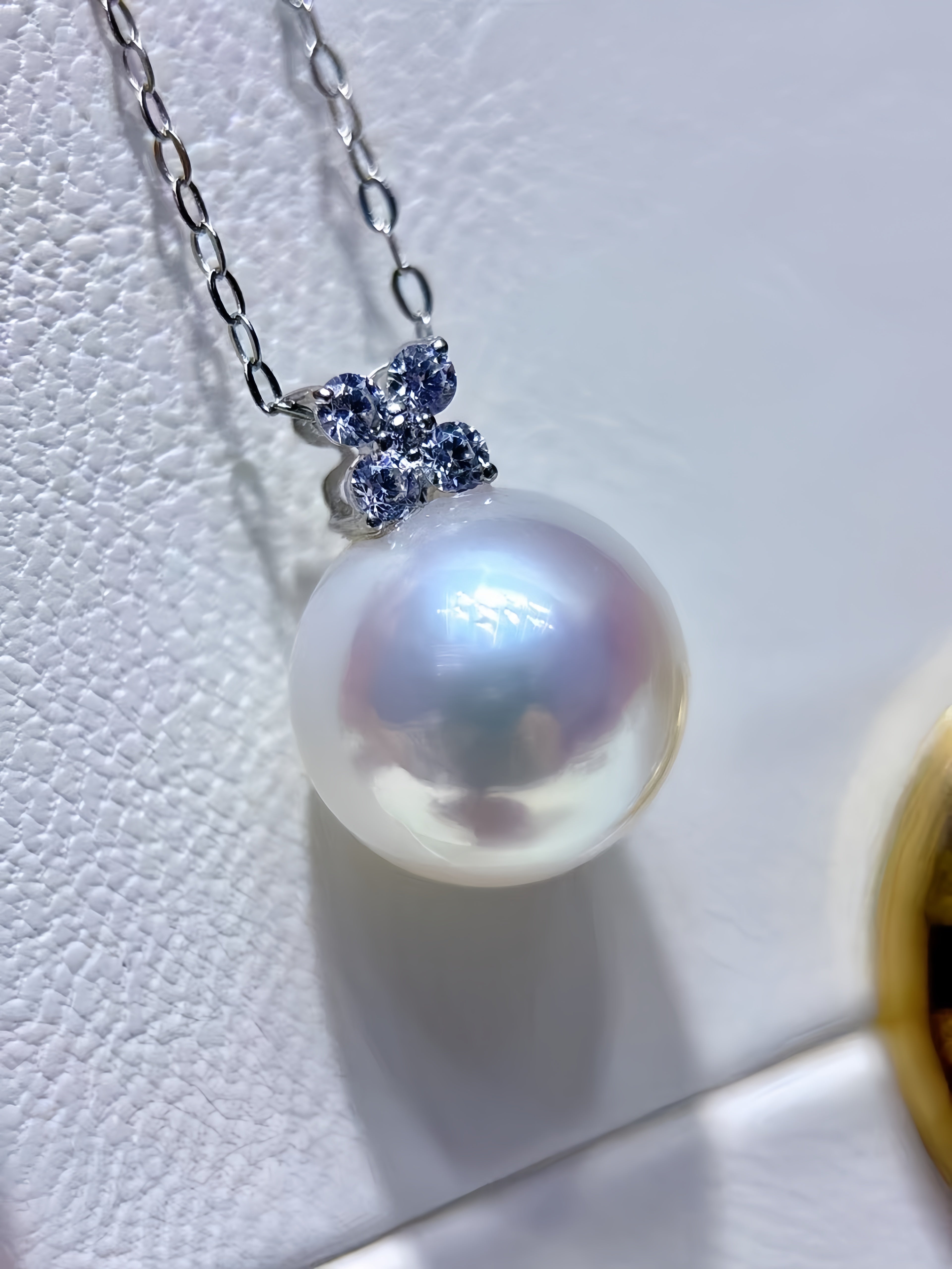 A close-up of a silver chain necklace with a large, lustrous white pearl pendant. The pendant is accented by a small four-stone floral cluster set with dark blue gemstones, resting on a textured white surface.