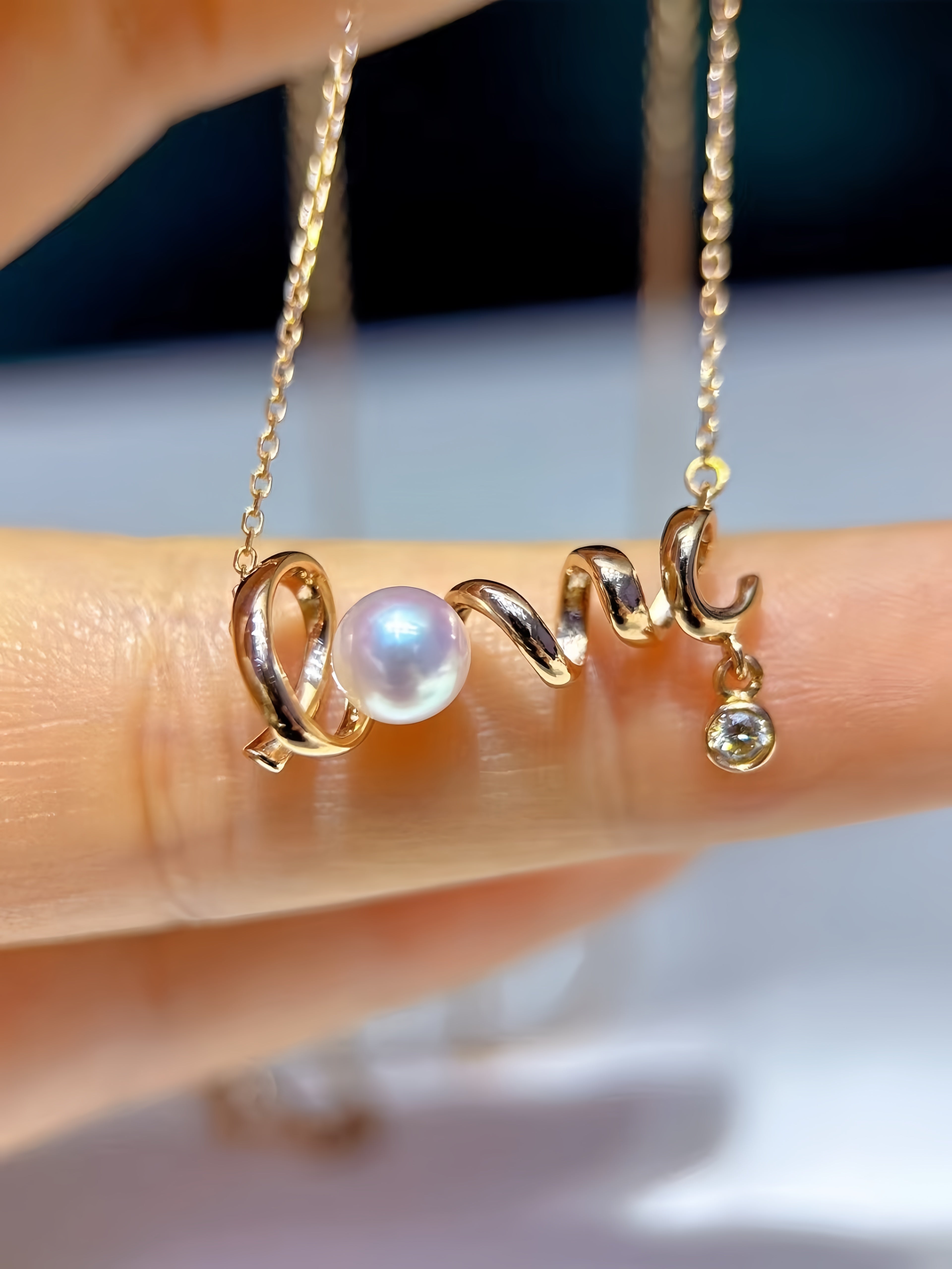 A close-up of a gold chain necklace being held between fingers. The pendant features a curved gold design with a central white pearl and a small dangling clear gemstone.